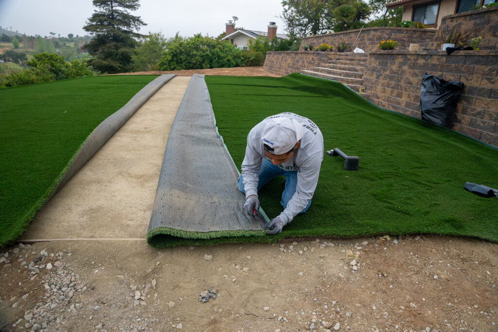 A man trims a the edge of a large turf sheet to fit against the one beside it